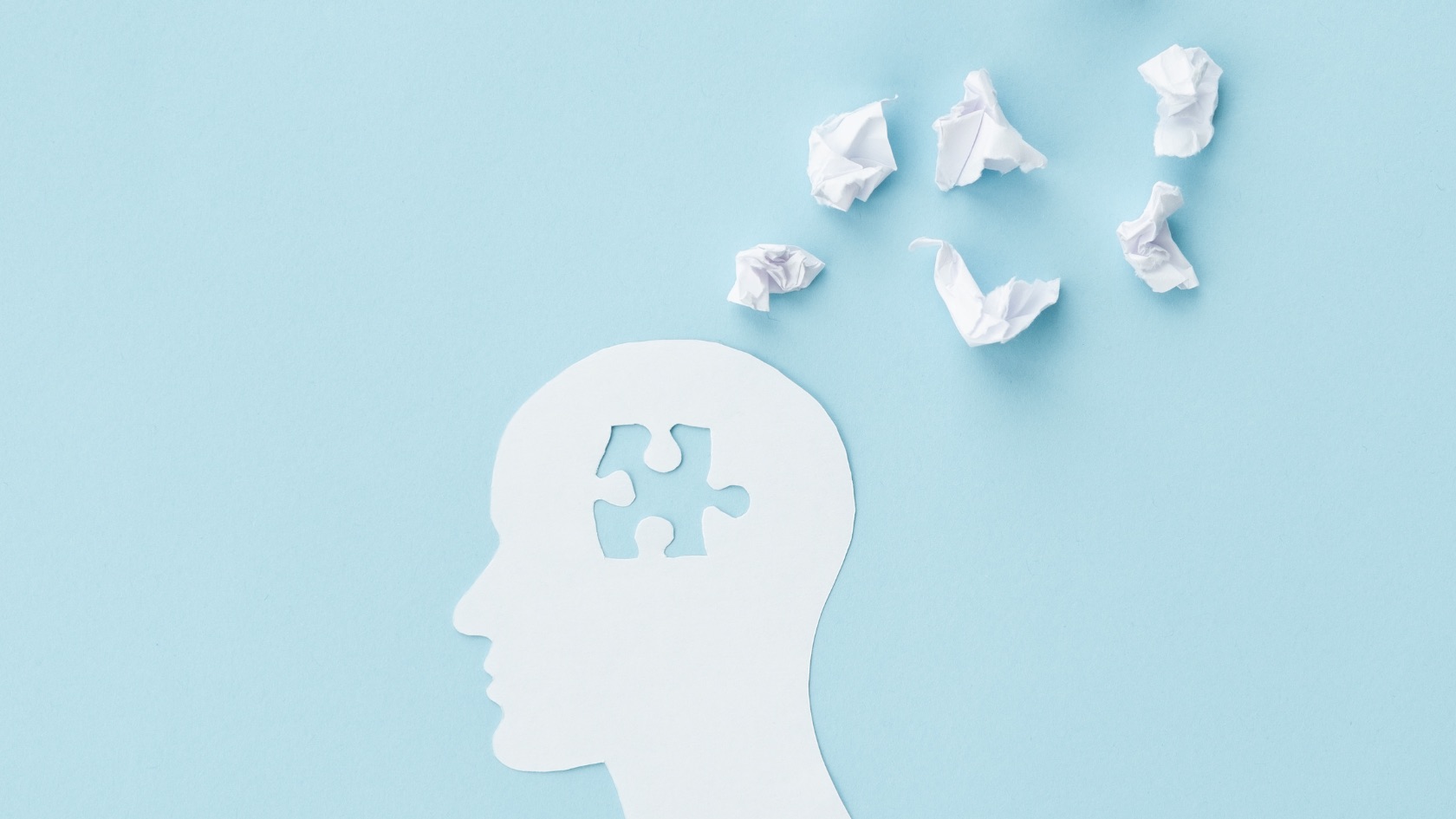 White paper cut-out profile of a head on a light blue background with a puzzle-piece shape cut out from the brain area, with small crumpled paper balls scattered above.