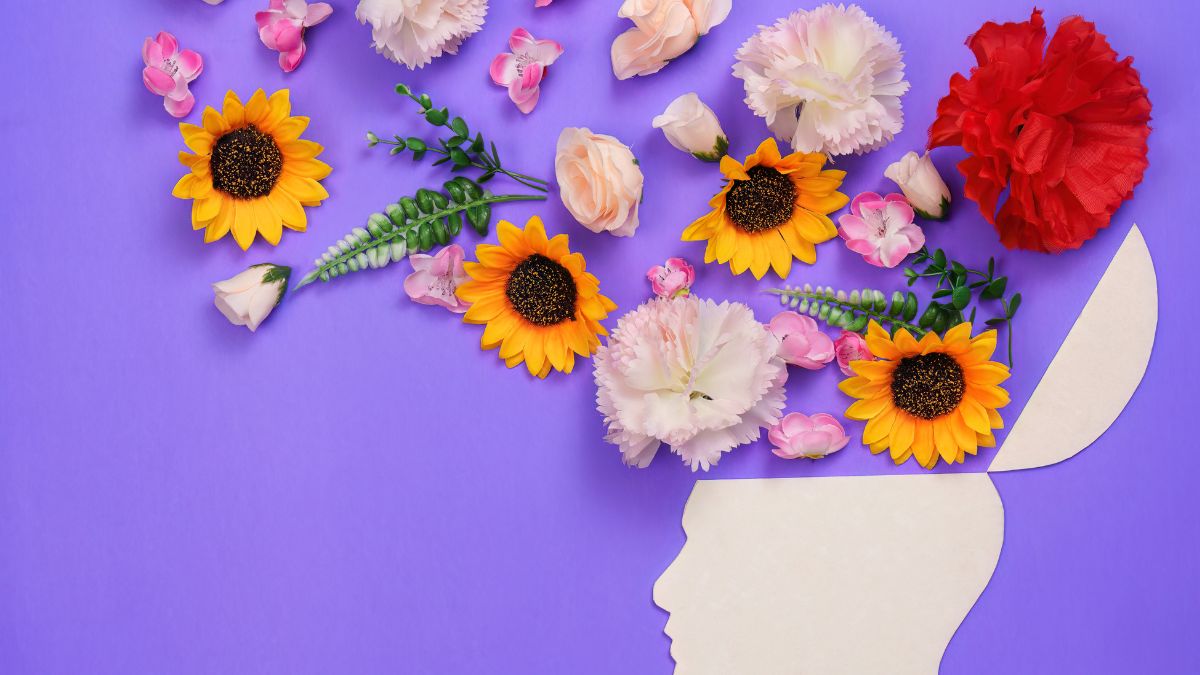 Flat lay of mixed flowers on a purple background, including yellow sunflowers, pale pink and white blossoms, and a red flower, with a white paper cut-out profile of a head in the bottom right.