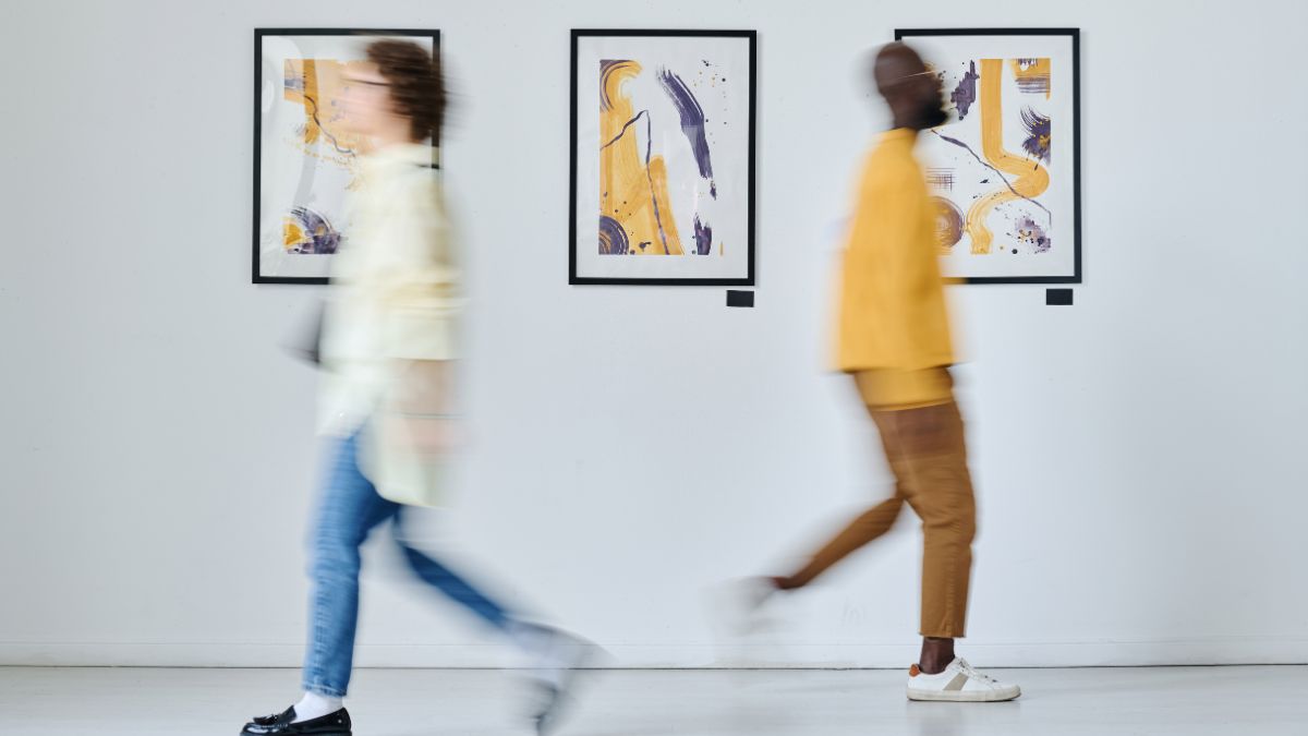 Featured image: two blurred visitors walking past three framed abstract artworks on a white gallery wall in a modern gallery setting.